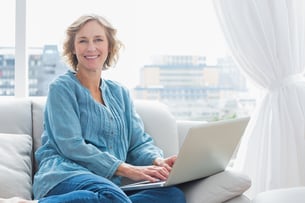 Content blonde woman sitting on her couch using laptop smiling at camera at home in the sitting room