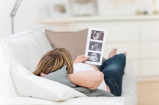 Rear view of pregnant woman holding babys ultrasound scan while lying on couch at home